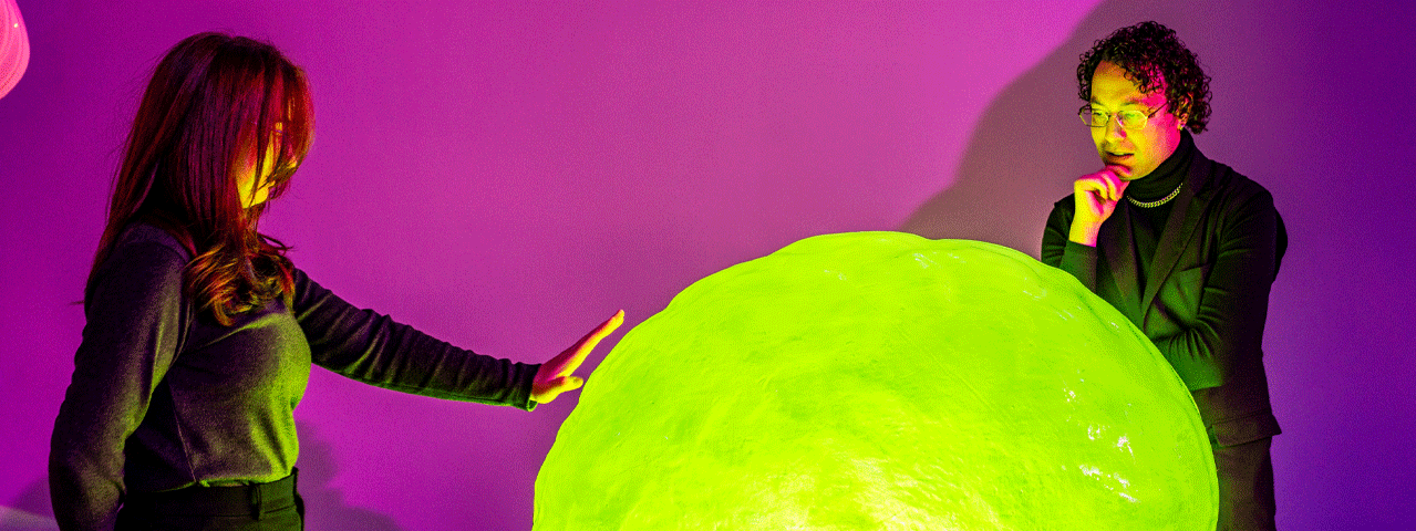 Twee bezoekers bewonderen een groot, groen lichtgevend object dat ze aanraken. Bezoek Nxt Nature Museum Gratis met de VIP-KAART!