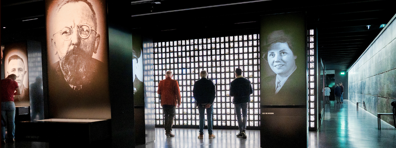 Drie bezoekers bekijken de tentoonstelling in Nationaal Monument Kamp Amersfoort, in een donkere ruimte met oplichtende foto's. Deelnemers bezoeken dit monument gratis met de VIP-KAART.