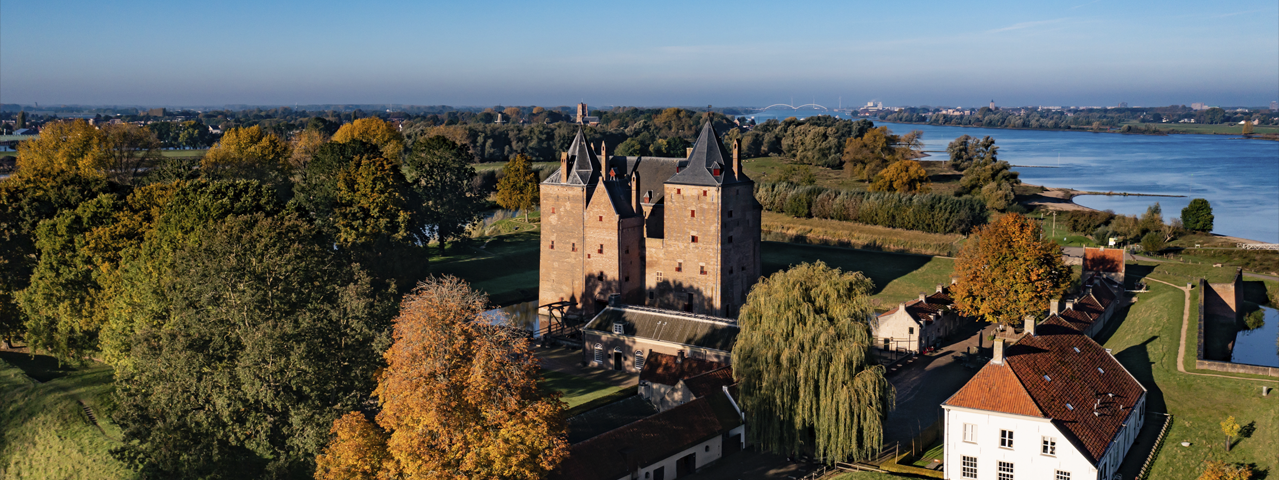 Een prachtig uitzicht op Kasteel Loevestein, omringd door groene velden, kleurrijke bomen en waterpartijen. De robuuste torens en muren van het kasteel steken imposant af tegen de heldere blauwe lucht. VIP-KAART-houders genieten van gratis entree tot dit iconische kasteel en de bijbehorende tentoonstellingen.