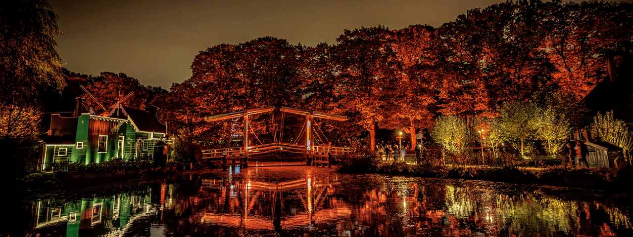 Een betoverend verlichte avondscène in het Openluchtmuseum, met een ophaalbrug, molen en bomen die warm oplichten in herfstkleuren, prachtig weerspiegeld in het water. VIP-KAART-houders genieten van gratis toegang tot dit sfeervolle museum.