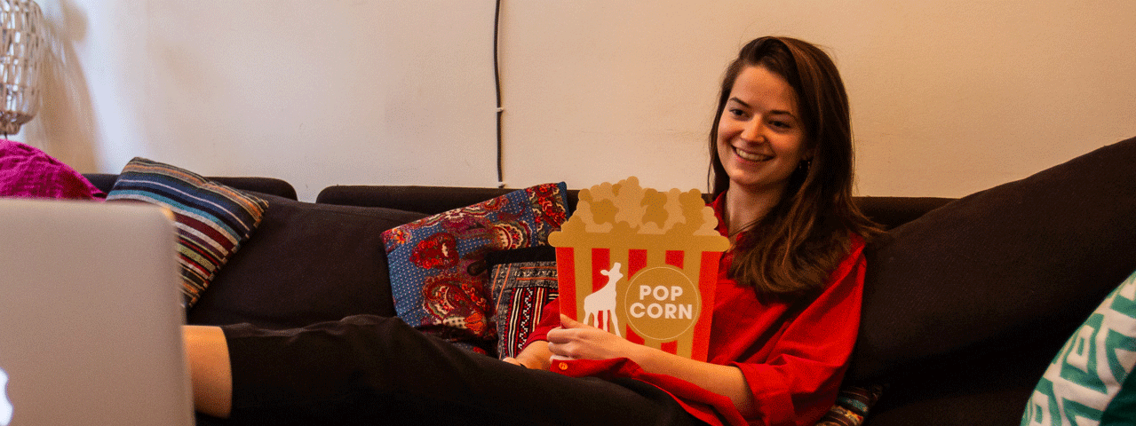 Een vrouw kijkt naar een film op haar laptop vanaf de bank. In haar handen draagt ze een kartonnen bord met een afbeelding van popcorn en het logo van het Nederlands Film Festival. Deelnemers krijgen korting op Pathé Thuis films tijdens het NFF.