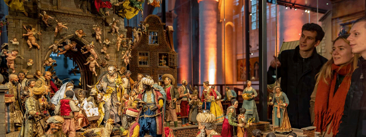 Bezoekers gekleed in winterse kleding bewonderen een rijk uitgevoerde, historische kerststal vol houten figuren.