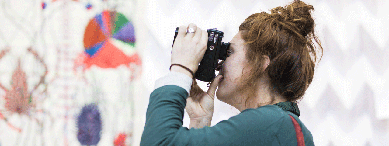 Een jonge vrouw met haar haar in een knot maakt met een fotocamera een foto van een kleurrijk kunstwerk in Textielmuseum Tilburg. Een bezoek aan het museum is gratis met de VIP-KAART.