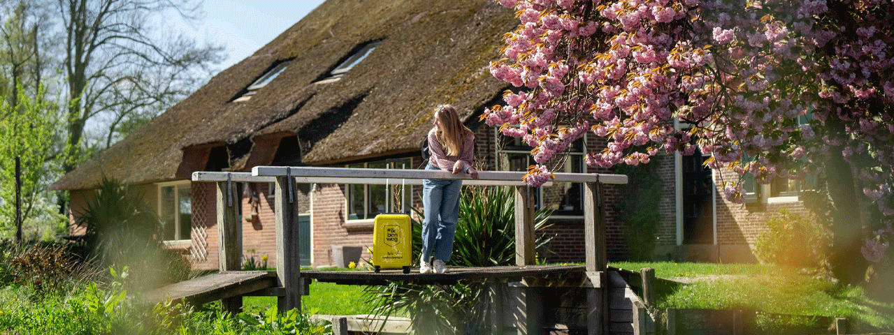Een vrouw leunt tegen een houten brug op een zonnige dag bij een kersenbloesem en een oude boerderij in Giethoorn.