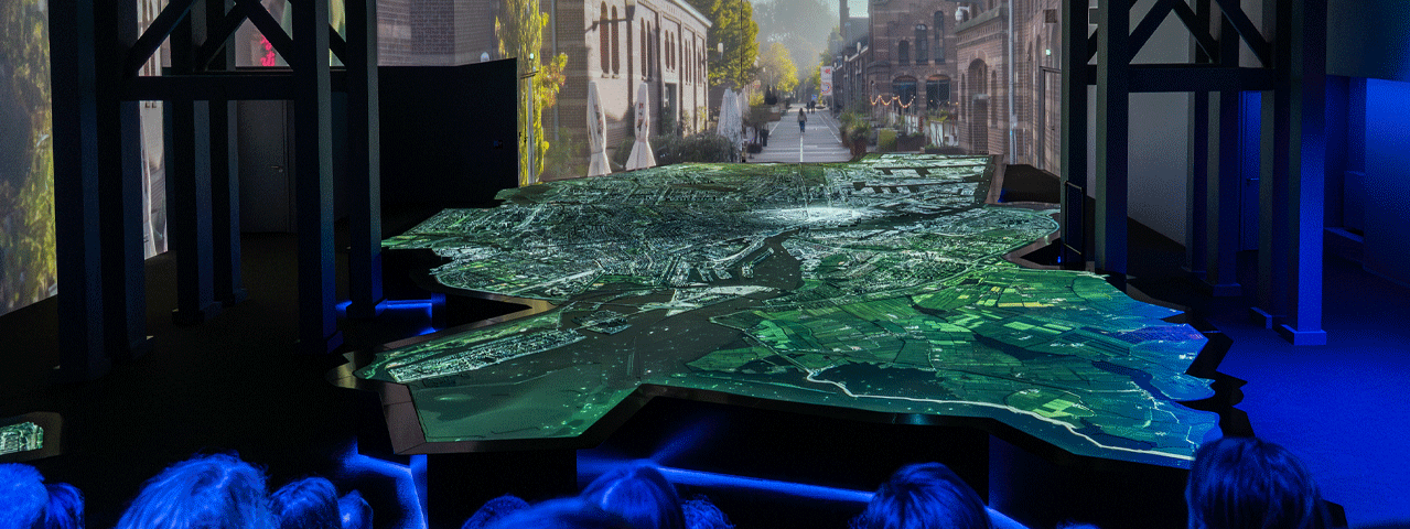 Een in blauw licht badend publiek kijkt naar een dynamische projectie op een grote maquette van de stad Amsterdam. Deze experience is gratis te bezoeken met de VIP-KAART.