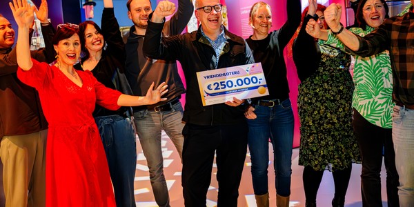 Een groep enthousiaste mensen viert samen een donatie van de VriendenLoterij. In het midden houdt een man lachend een groot bord omhoog met daarop “VriendenLoterij €250.000,- Limburgs Museum”.
