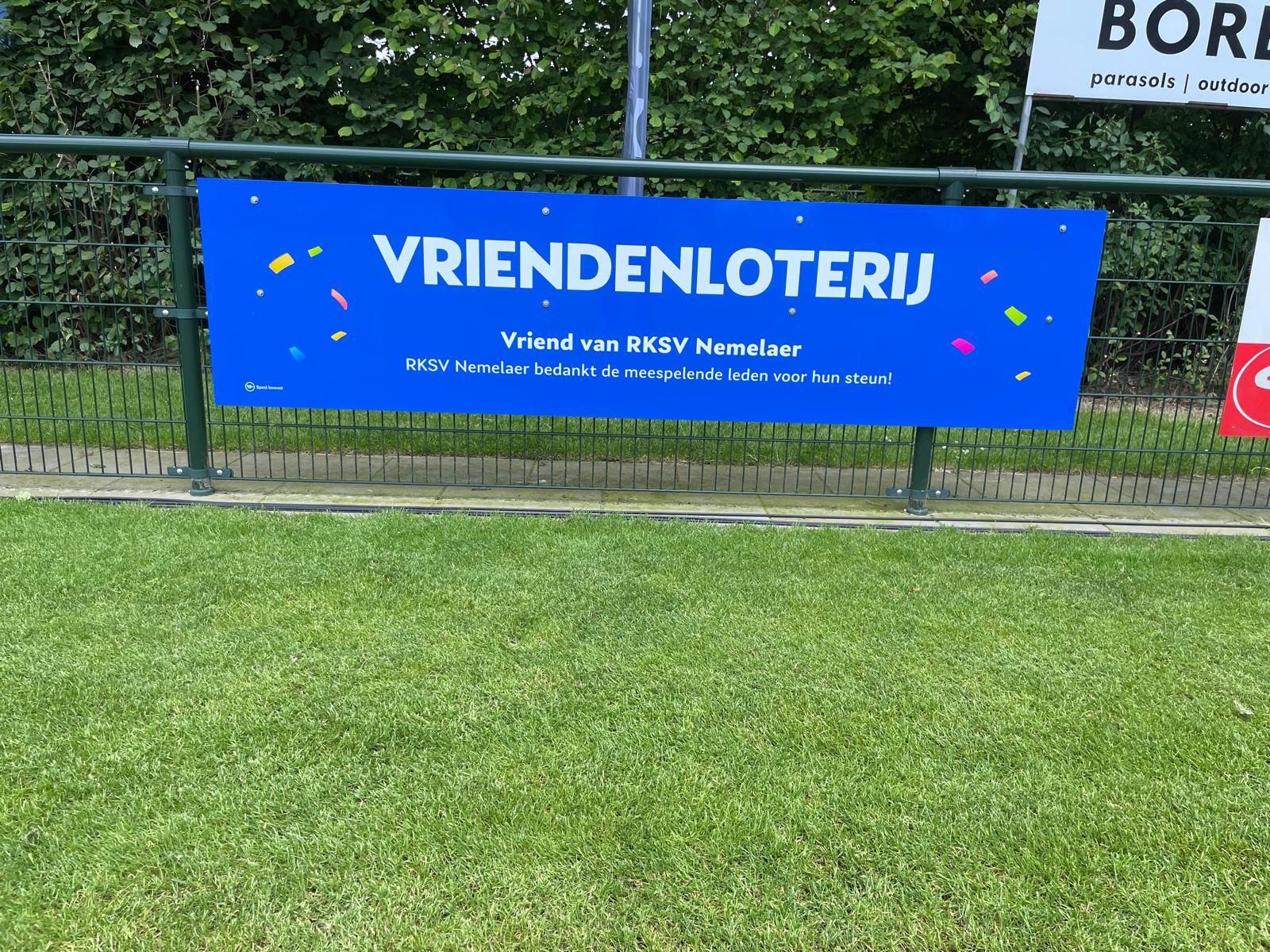Blauw bord met tekst ‘Vriendenloterij, Vriend van RKSV Nemelaer’ langs een grasveld.