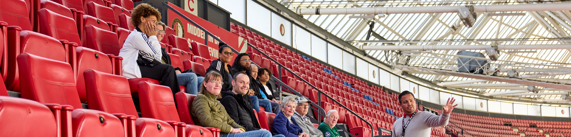 Groep bezoekers op een stadiontour in een groot voetbalstadion, zittend in rode stoelen met een gids die uitleg geeft. Het stadion is leeg en baadt in natuurlijk licht, wat de uitgestrekte grootte en structuur benadrukt.