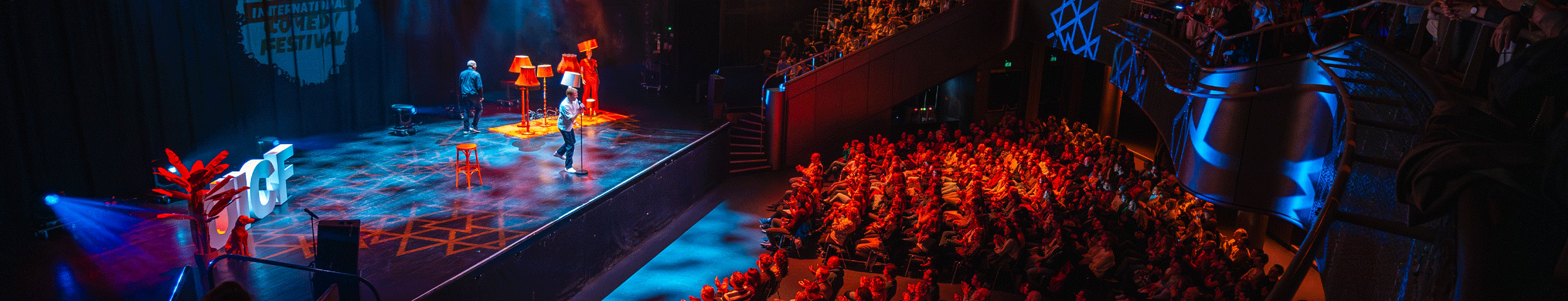 Zicht op een volle theaterzaal, waarin het publiek naar een stand-up comedian kijkt op een podium versierd met lampen. De zaal baadt in rood en blauw licht. Ontvang korting op tickets voor het Utrecht International Comedy Festival met de VIP-KAART!
