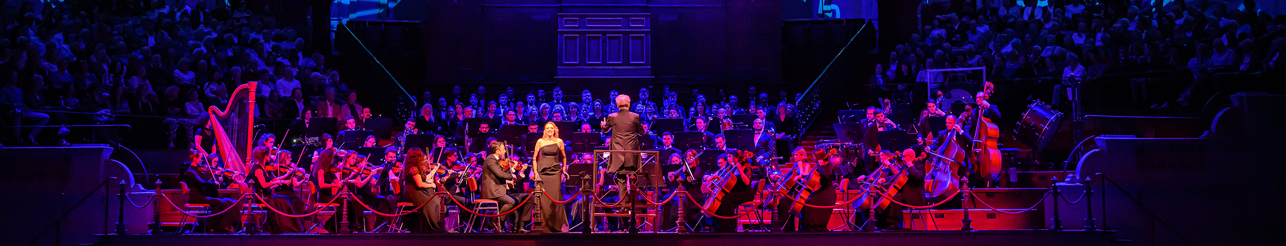 Een orkest met een zangeres in een zwarte jurk speelt in de grote zaal van Het Concertgebouw. De ruimte is verlicht met rode en blauwe decorlichten. Deelnemers krijgen korting op concerten met hun VIP-KAART.