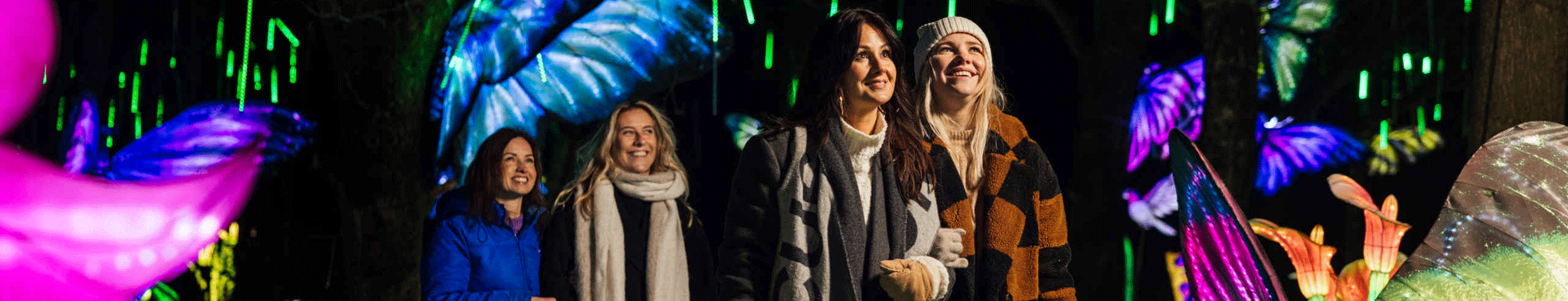 Vier vriendinnen in winterkleding lopen door een kleurrijke wereld vol lichtgevende kunstwerken.