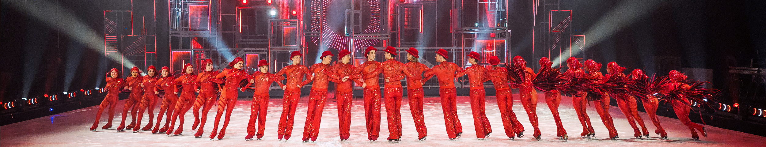 Een groep kunstschaatsers in rode kleding met glitters performen de show HORIZONS van Holiday on ICE. Deelnemers profiteren van korting op deze magische show met de VIP-KAART.