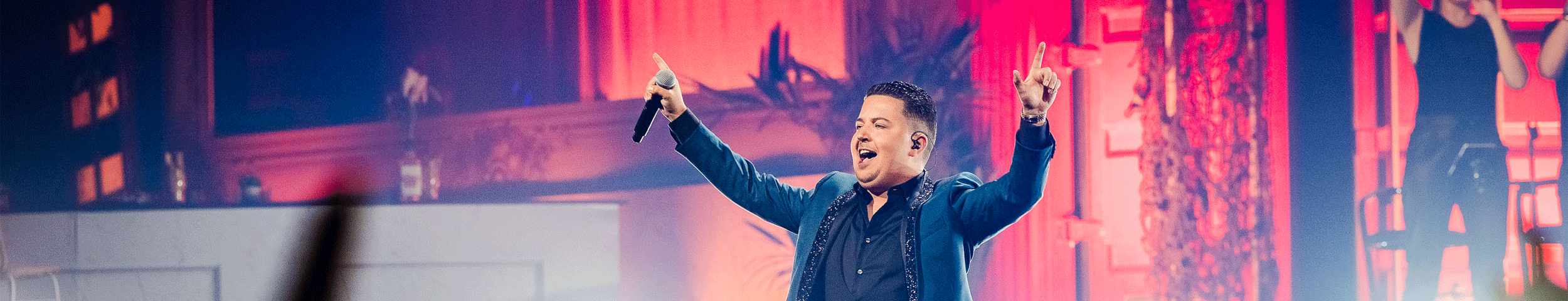 Een mannelijke zanger in een blauwe blazer treedt energiek op een kleurrijk podium op, met zijn armen omhoog, terwijl het publiek op de voorgrond juicht. Ontvang 50% korting op ticketgs voor Dikste Cafe aand e haven met de VIP-KAART!