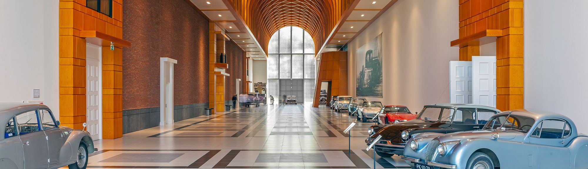 De indrukwekkende, lichte hal van het Louwman Museum. Aan de zijkant van de hal staan oude auto's en oldtimers geparkeerd. Aan het einde van de hal verlicht een groot raam dat tot het plafond reikt de hele hal.