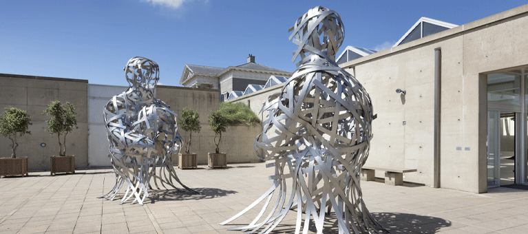 Twee grote abstracte mensfiguren gemaakt van verweven metalen strips staan tegenover elkaar in een zonnige, openlucht patio met jonge boompjes en moderne architectuur — dit is een gratis toegankelijke tentoonstelling in Museum Beelden aan Zee met de VIP-KAART.