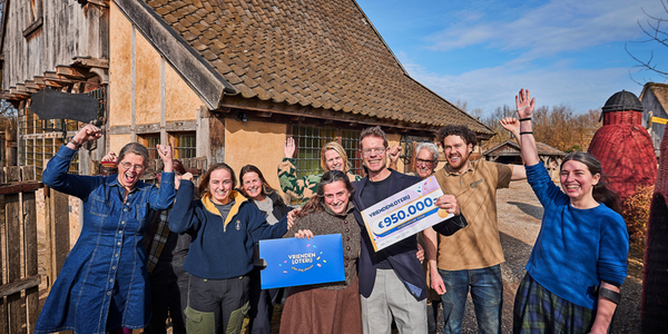 Een groep medewerkers van Museumpark Vonk viert samen een schenking van de VriendenLoterij. Ze poseren lachend met een cheque van €950.000 voor een historisch gebouw op het terrein.