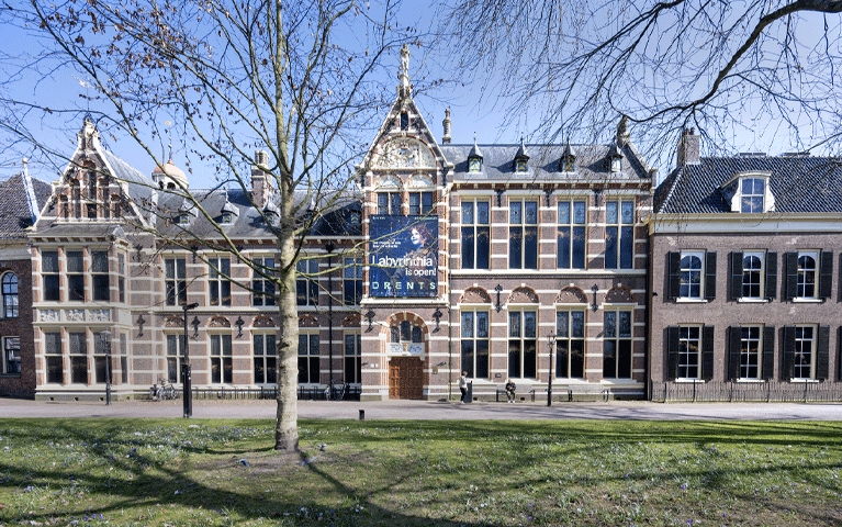 Het vooraanzicht van het historische, bakstenen gebouw van het Drents Museum. Op de voorgrond is een grasveld met bomen te zien. Boven de deur hangt een grote poster over de tentoonstelling Labyrinthia. Ontdek het Drents Museum gratis met uw VIP-KAART!