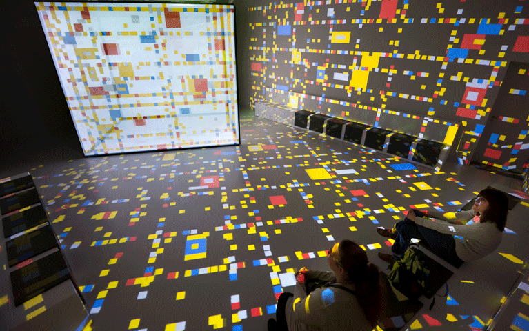 Bezoekers zitten in een kleurrijke projectieruimte in het Mondriaanhuis, omringd door bewegende patronen van primaire kleuren en geometrische vormen die doen denken aan Mondriaans kenmerkende stijl. De immersieve installatie laat de wereld van Mondriaan op een moderne en interactieve manier tot leven komen.