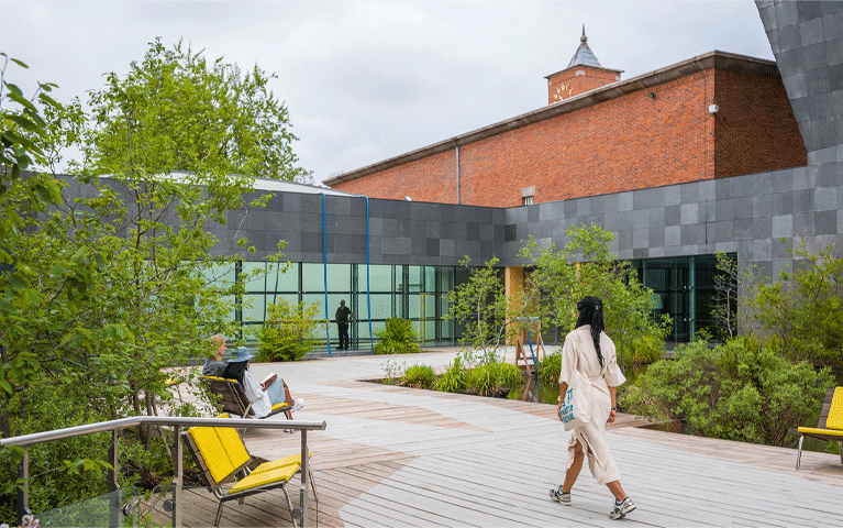 Mensen kuieren en lopen rond in de binnentuin van een museum, dat bestaat uit een laag, grijs gebouw en een hoger, bakstenen gebouw met toren. Ontdek het Van Abbemuseum helemaal gratis met de VIP-KAART!