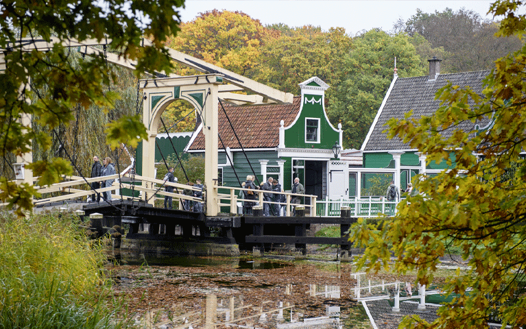 Bezoekers lopen over een historische brug en langs authentieke Zaanse huisjes tussen de bossen bij het Nederlands Openluchtmuseum. Ontvang gratis entree bij het Nederlands Openluchtmuseum met de VIP-KAART!