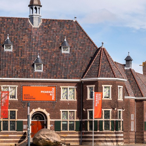 Historisch pand van Rijksmuseum Twenthe in Enschede, met oranje banners voor een Picasso & Matisse-tentoonstelling. Het gebouw heeft een klassiek pannendak en glas-in-loodramen. Bezoekers met een VriendenLoterij VIP-KAART krijgen hier gratis toegang.