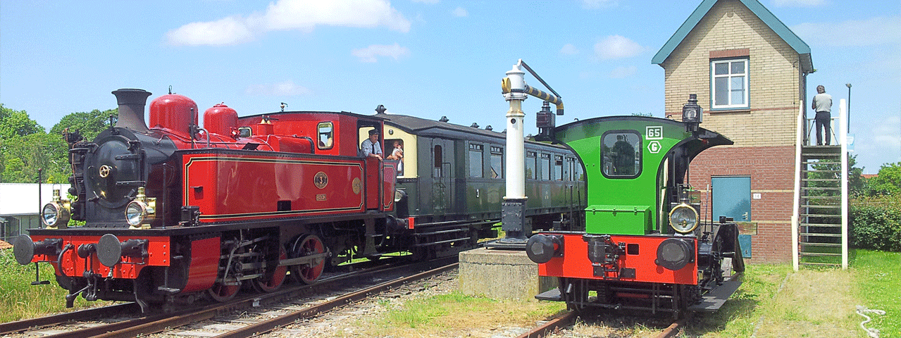 Vooraanzicht van de locomotief van de historische stoomtrein Goes-Borsele, naast een wachtershuisje op een zonnige dag met blauwe lucht. Met de VIP-KAART reis je met extra voordeel mee op deze historische stoomtrein.