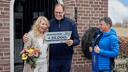 Een stel bekijkt blij een cheque van €25.000 van de VriendenLoterij terwijl Wolter Kroes toekijkt.