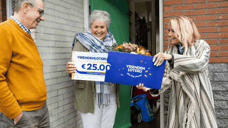 Een vrouw ontvangt aan haar voordeur een cheque van €25.000 van de VriendenLoterij en een bos bloemen.