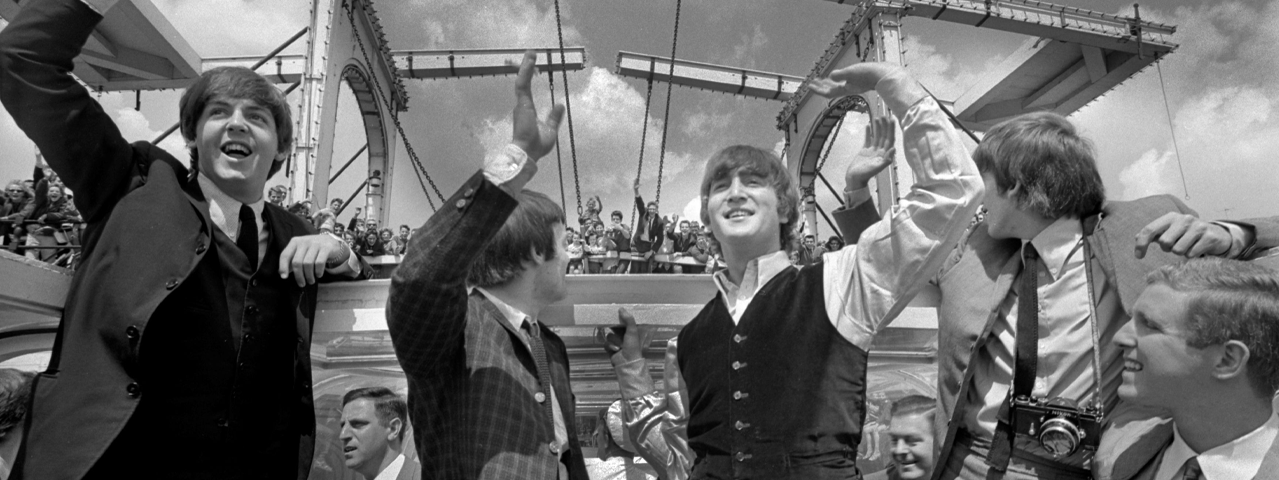 Zwart-wit foto van de beatles in formele kleding die zwaaien naar de toeschouwers boven hen op de brug
