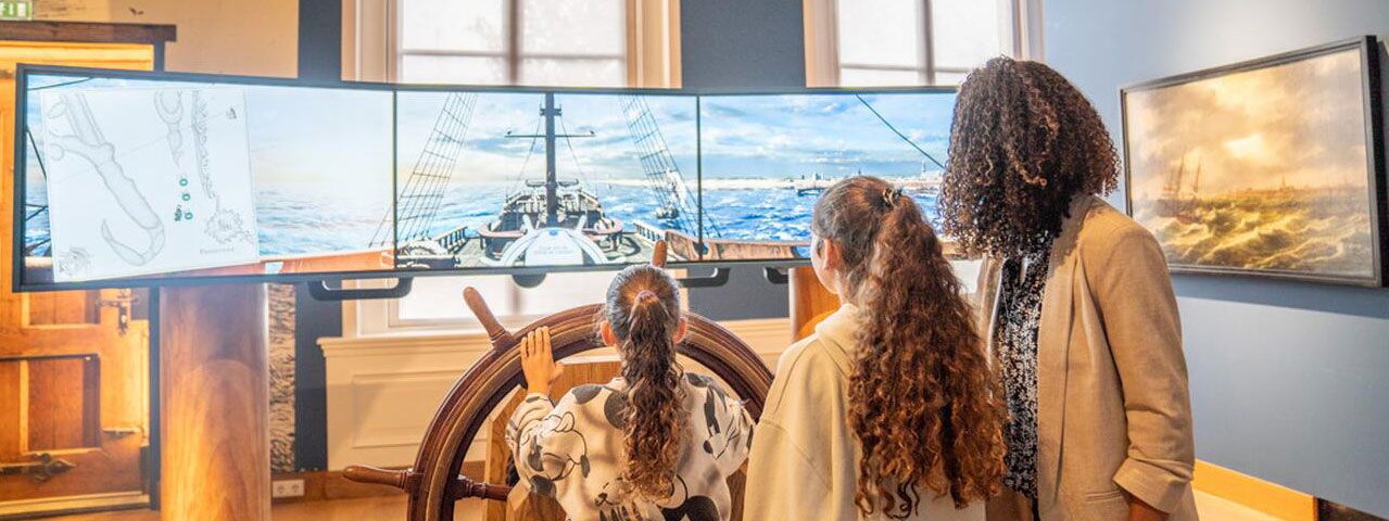 Een vrouw en twee kinderen kijken naar een simulatie van een schip op drie grote schermen terwijl het jongste kind aan een groot stuur draait.