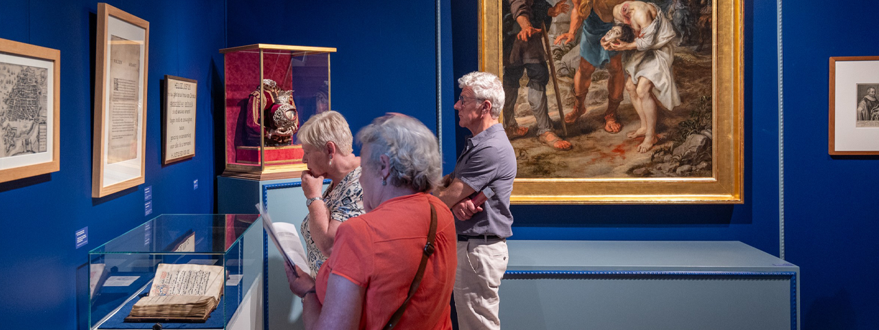Drie oudere bezoekers bekijken geconcentreerd een oud document in een blauwe museumzaal gevuld met schilderijen en andere objecten