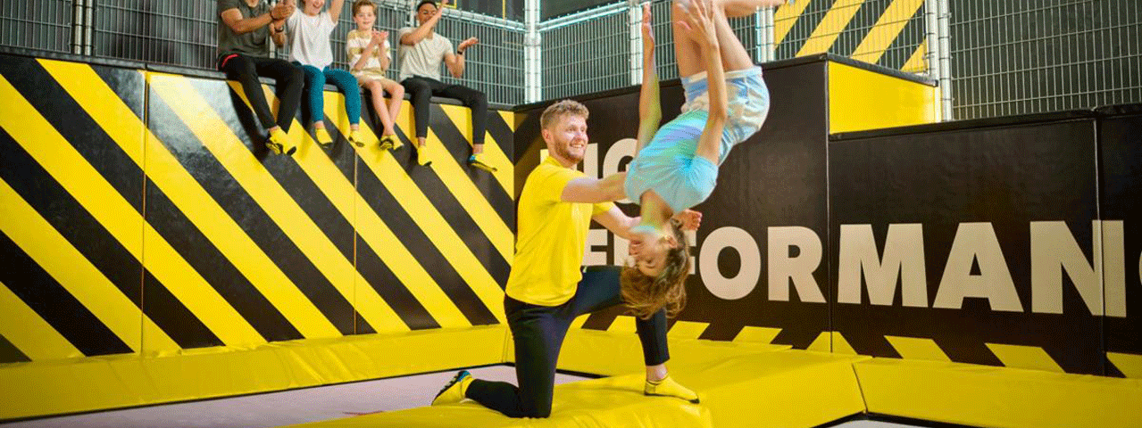 Een man helpt een vrouw met het maken van een salto op een trampoline in een zwart-geel versierde ruimte van You Jump.
