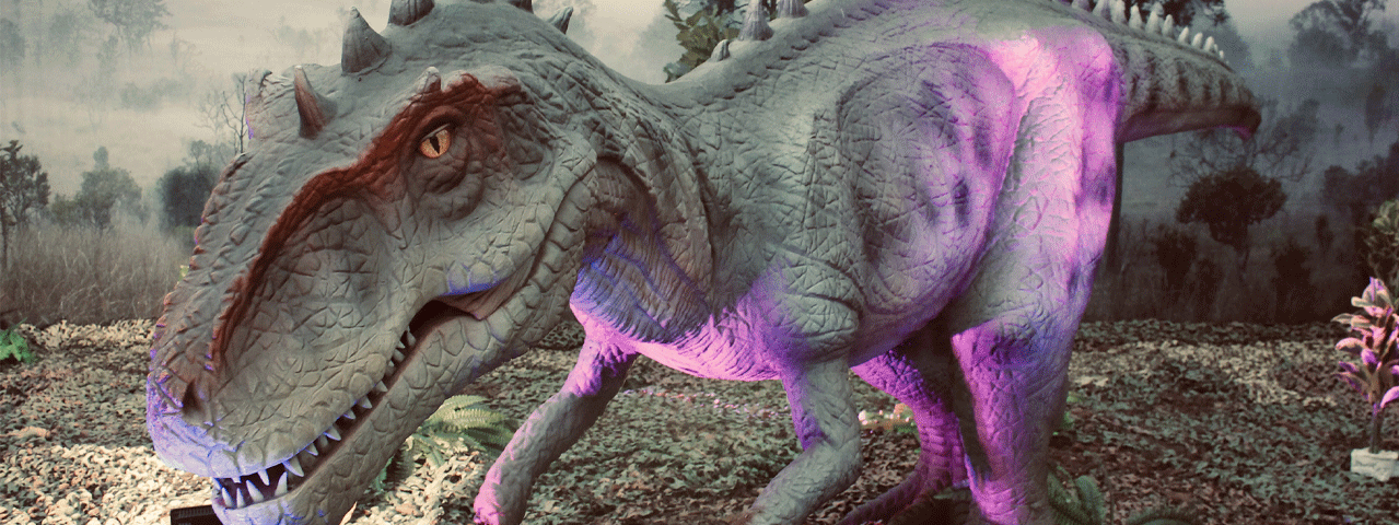 Een grijze dinosaurus in een bos bij World of Dinos. Een leuk uitje met korting met de VIP-KAART