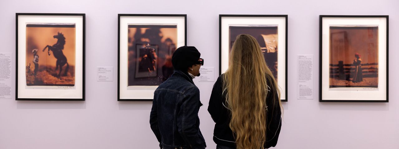 Twee bezoekers kijken geïnteresseerd naar vier foto's in zwarte lijsten in het H'ART Museum.