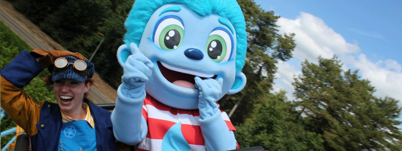 Een vrouw houdt lachend haar pet met bril vastnaast een grote blauwe mascotte in de achtbaan bij Dippie Doe