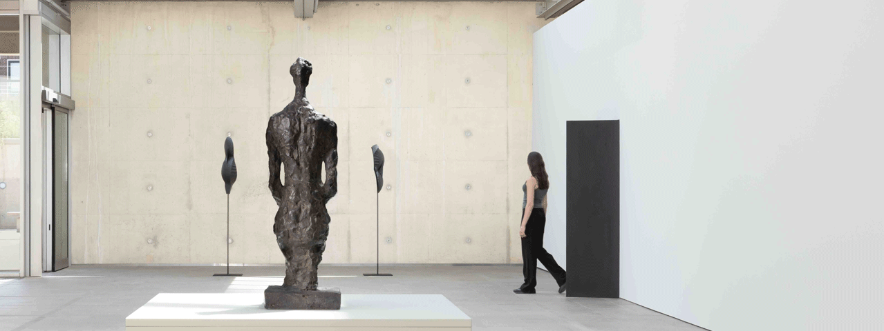 Een vrouw loopt via een deur een minimalistische tentoonstellingsruimte binnen met drie sculpturen