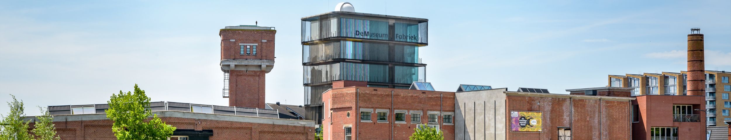 Een bakstenen industrieel gebouwencomplex op een zonnige dag met op de voorgrond een vijver met een loopbrug en gras. Ontdek De Museumfabriek gratis met de VIP-KAART