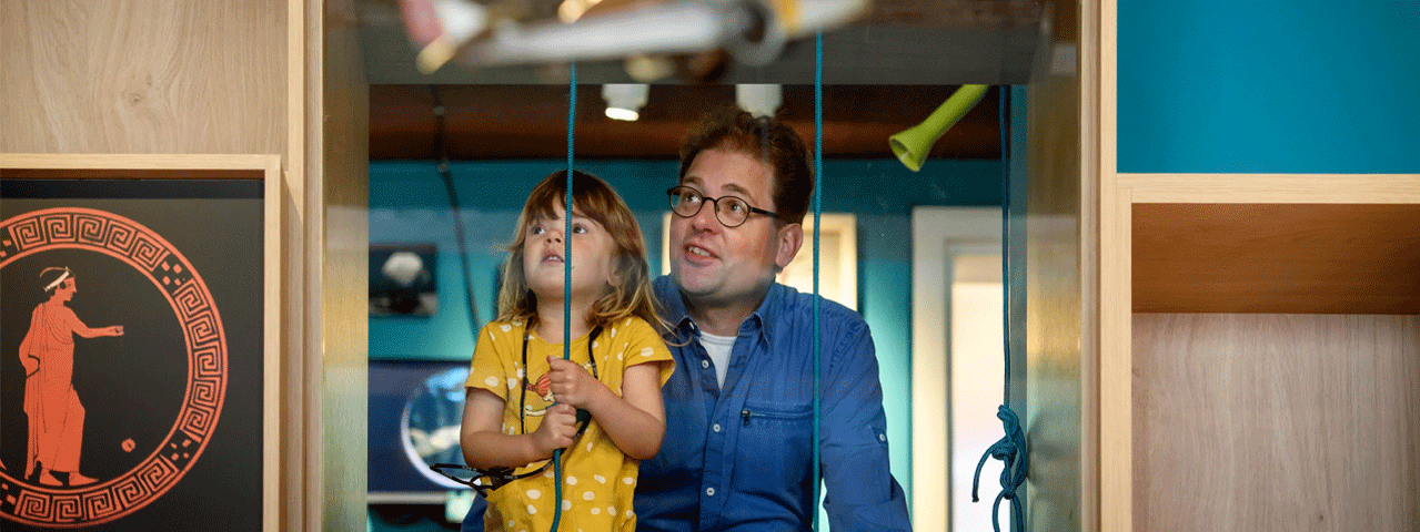 Een man en een kind kijken in een museum door een raam naar een interactieve opstelling. Bezoek het Speelgoedmuseum Deventer en ontvang er gratis entree met de VIP-KAART.
