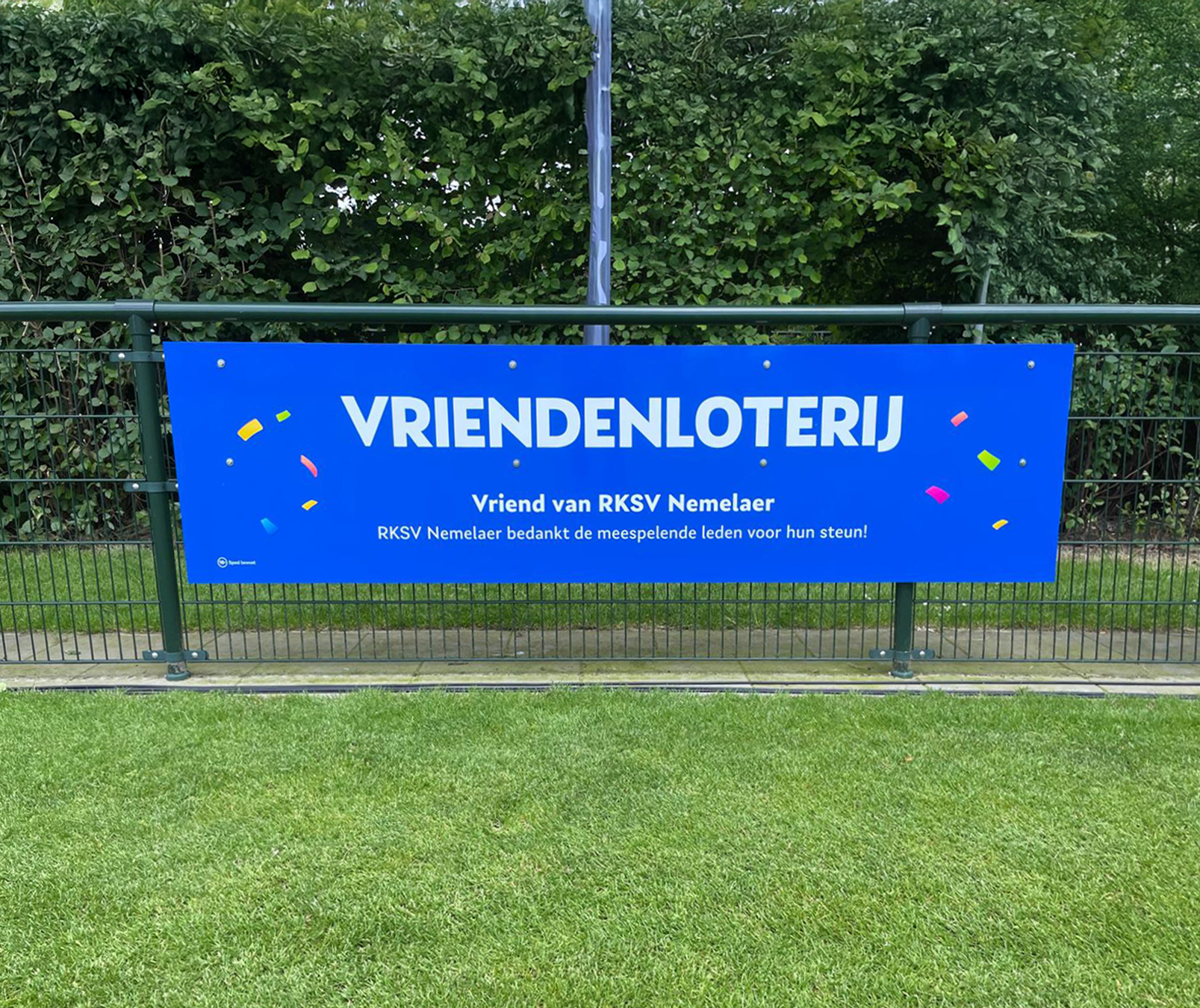 Blauw spandoek van de VriendenLoterij aan een hek bij RKSV Nemelaer, met confetti en dankwoord aan leden.