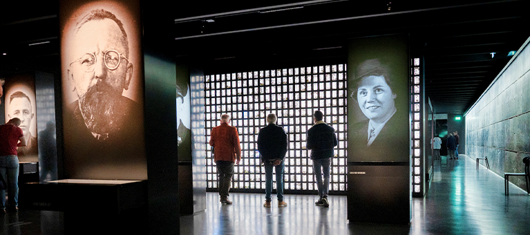 Drie bezoekers bekijken de tentoonstelling in Nationaal Monument Kamp Amersfoort, in een donkere ruimte met oplichtende foto's. Deelnemers bezoeken dit monument gratis met de VIP-KAART.