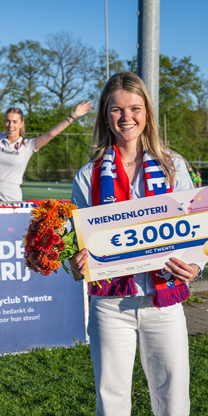 Een lid van HC Twente houdt een cheque van €3.000 van de VriendenLoterij vast, terwijl teamgenoten op de achtergrond juichen langs het hockeyveld.