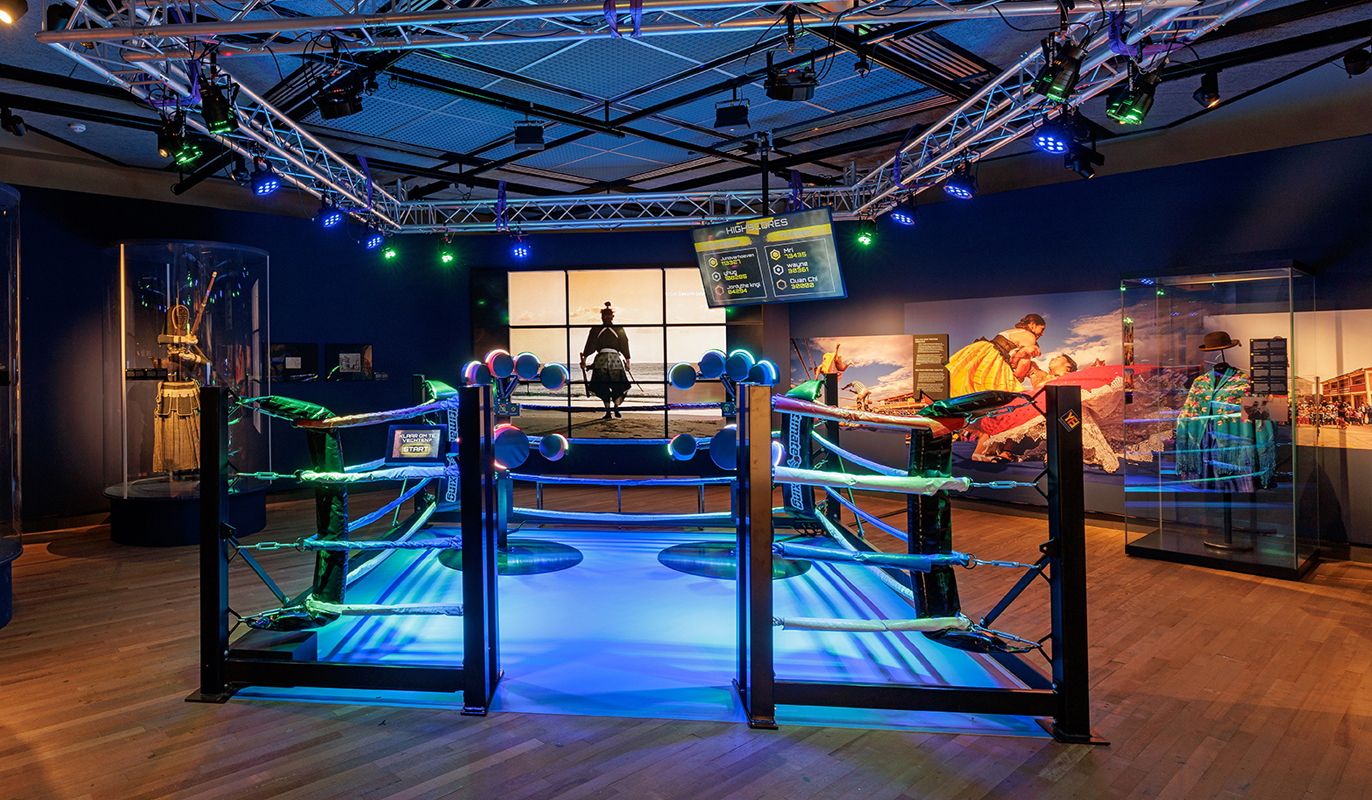 Zaalfoto van de tentoonstelling Martial Arts in Wereldmuseum Amsterdam. Middenin de zaal staat een grote boksring, omringd door historische kostuums.