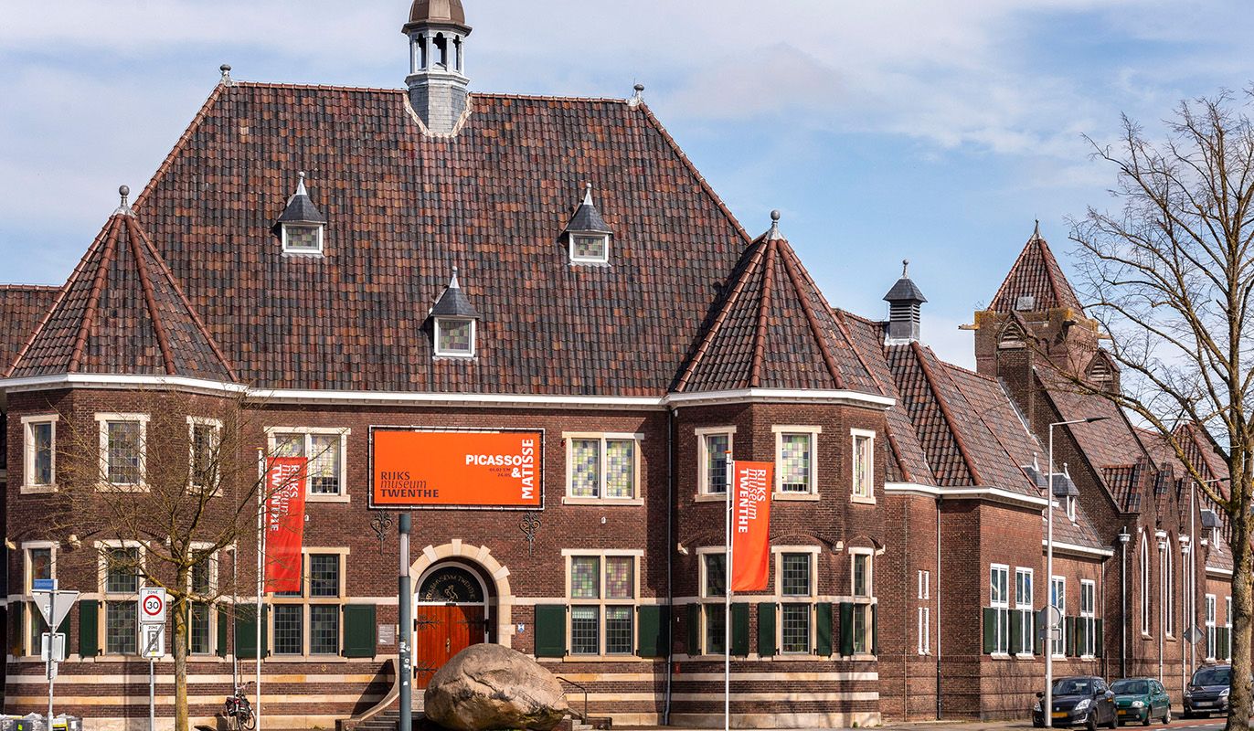 Historisch pand van Rijksmuseum Twenthe in Enschede, met oranje banners voor een Picasso & Matisse-tentoonstelling. Het gebouw heeft een klassiek pannendak en glas-in-loodramen. Bezoekers met een VriendenLoterij VIP-KAART krijgen hier gratis toegang.