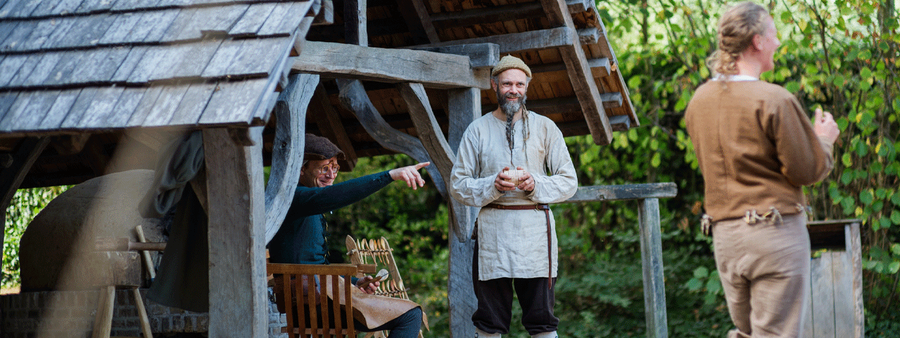 Drie mannen in Middeleeuwse kleding lachen en staan samen rondom een historische schuur in de openlucht. Bezoek het Prehistorisch Museum gratis met de VRiendenLoterij VIP-KAART!