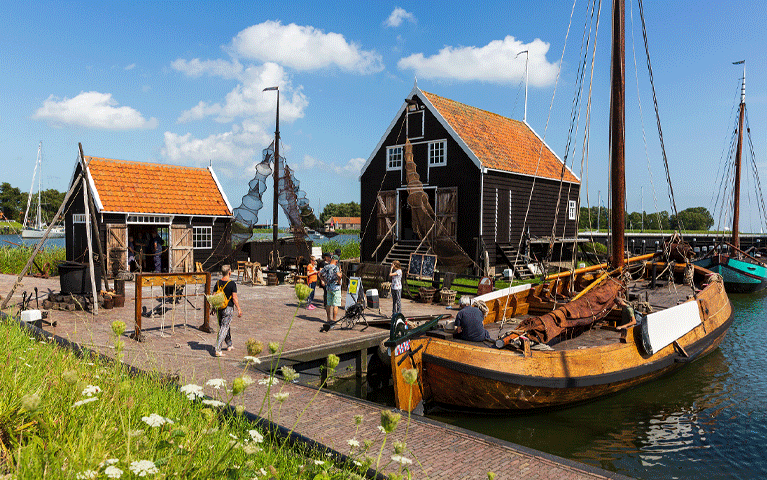 Een historische platbodem ligt aangemeerd bij een een typische vissershuisjes bij zonnig weer in het Zuiderzeemuseum. Het zuiderzeemuseum in Enkhuizen bezoekt u gratis met de VIP-KAART.i