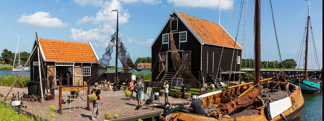 Een historische platbodem ligt aangemeerd bij een een typische vissershuisjes bij zonnig weer in het Zuiderzeemuseum. Het zuiderzeemuseum in Enkhuizen bezoekt u gratis met de VIP-KAART.i