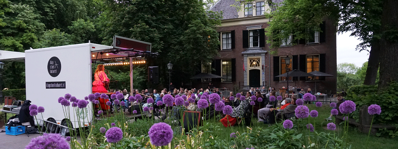 Voor een historische villa speelt een cabaretier in een felroze outfit op een mobiel podium (Klapstoelkabaret) voor een aandachtig publiek in tuinstoelen, omringd door paarse bloemen en groen — een sfeervol, gratis openluchtoptreden dat je met de VIP-KAART kunt bijwonen.