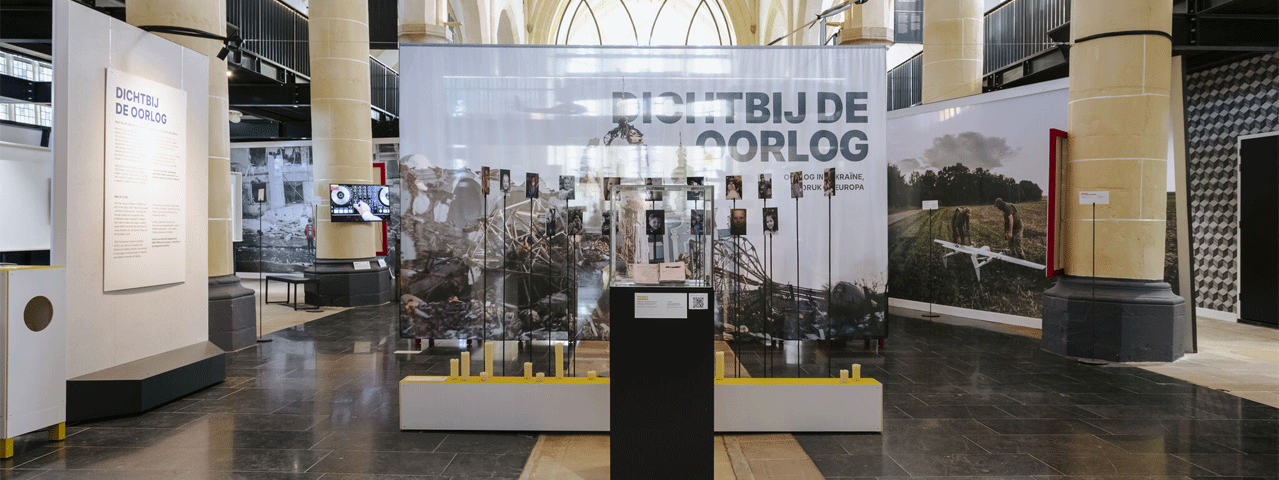 Zicht op een tentoonstelling met daarop een display en foto's in het thema van oorlog in een oude kerk met zuilen. Bezoek Dichtbij de oorlog in het Nationaal Museum Tachtigjarige Oorlog met de VIP-KAART gratis entree!