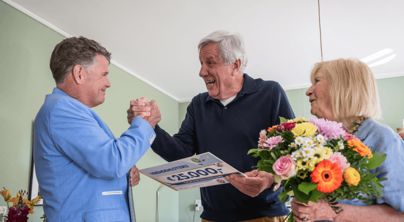 Een winnaar schudt Wolter Kroes de hand terwijl hij een cheque van €25.000 van de VriendenLoterij vasthoudt.