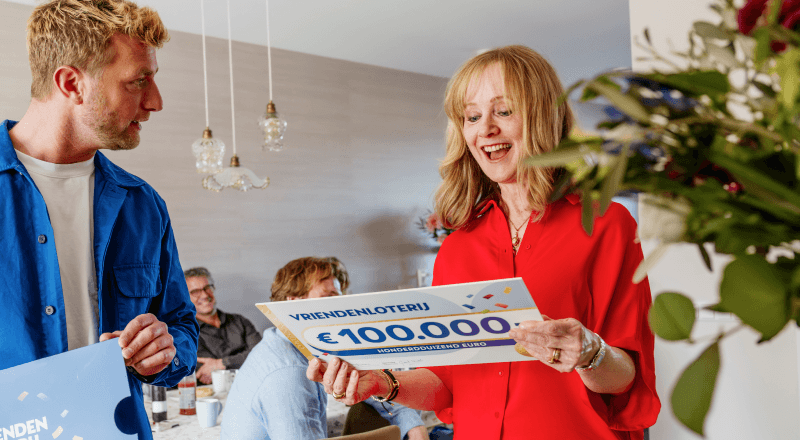 Een vrouw reageert blij op een cheque van €100.000 van de VriendenLoterij terwijl familie en vrienden toekijken.